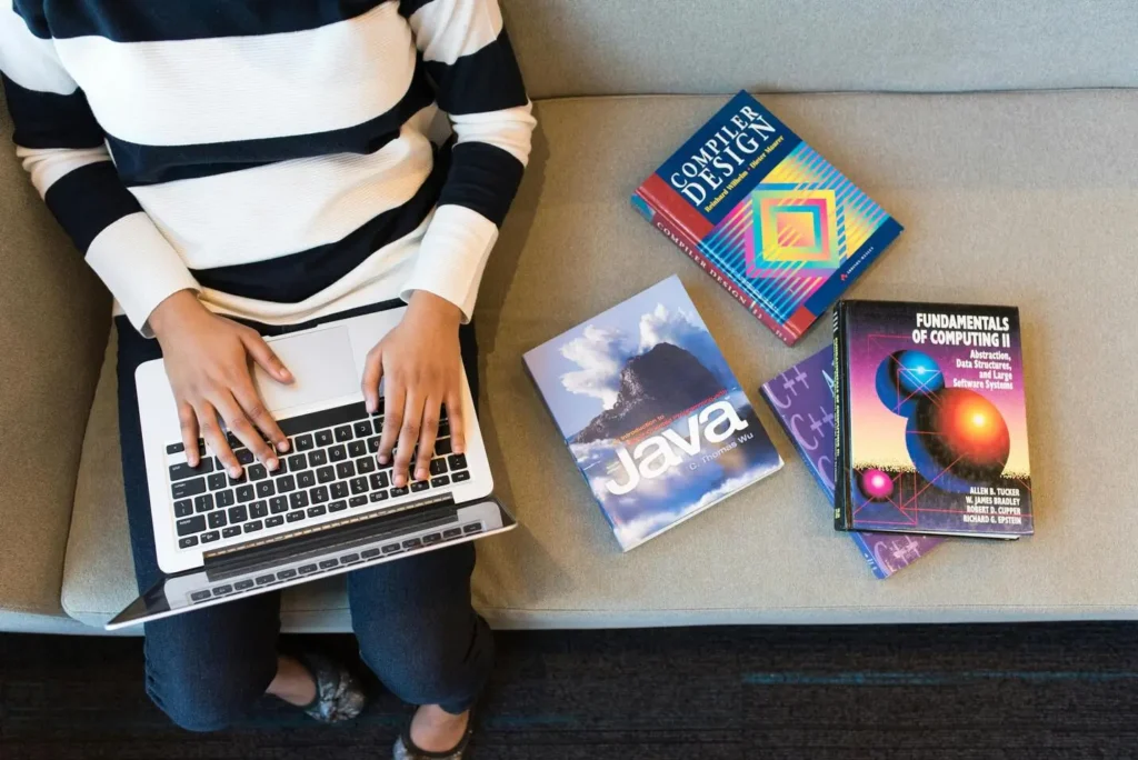 Mulher sentada em um sofá com notebook no colo e livros de design e programação ao lado.